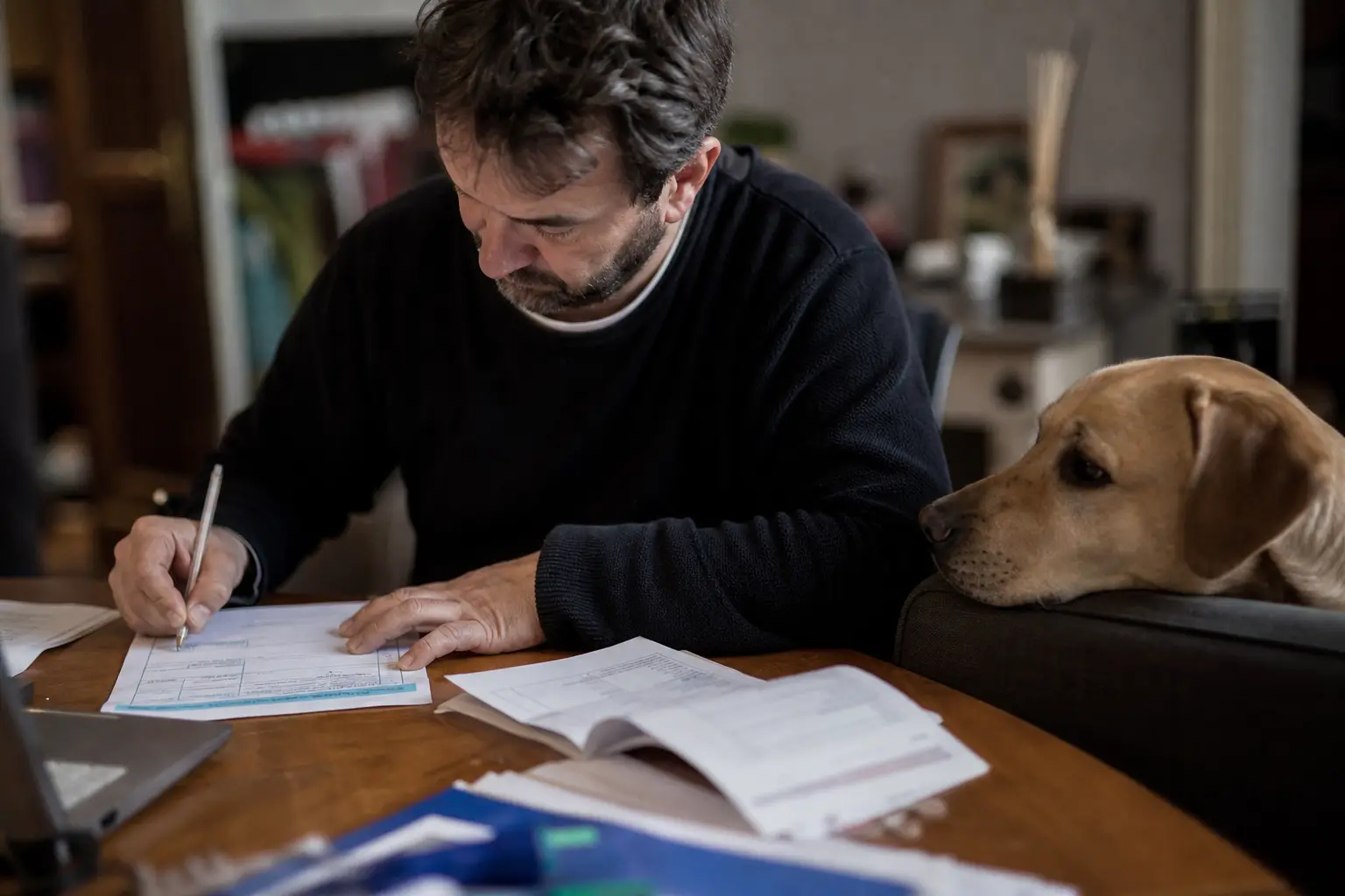 Homme remplissant un formulaire sur un bureau avec une facture à côté, son chien posant le menton sur l'accoudoir