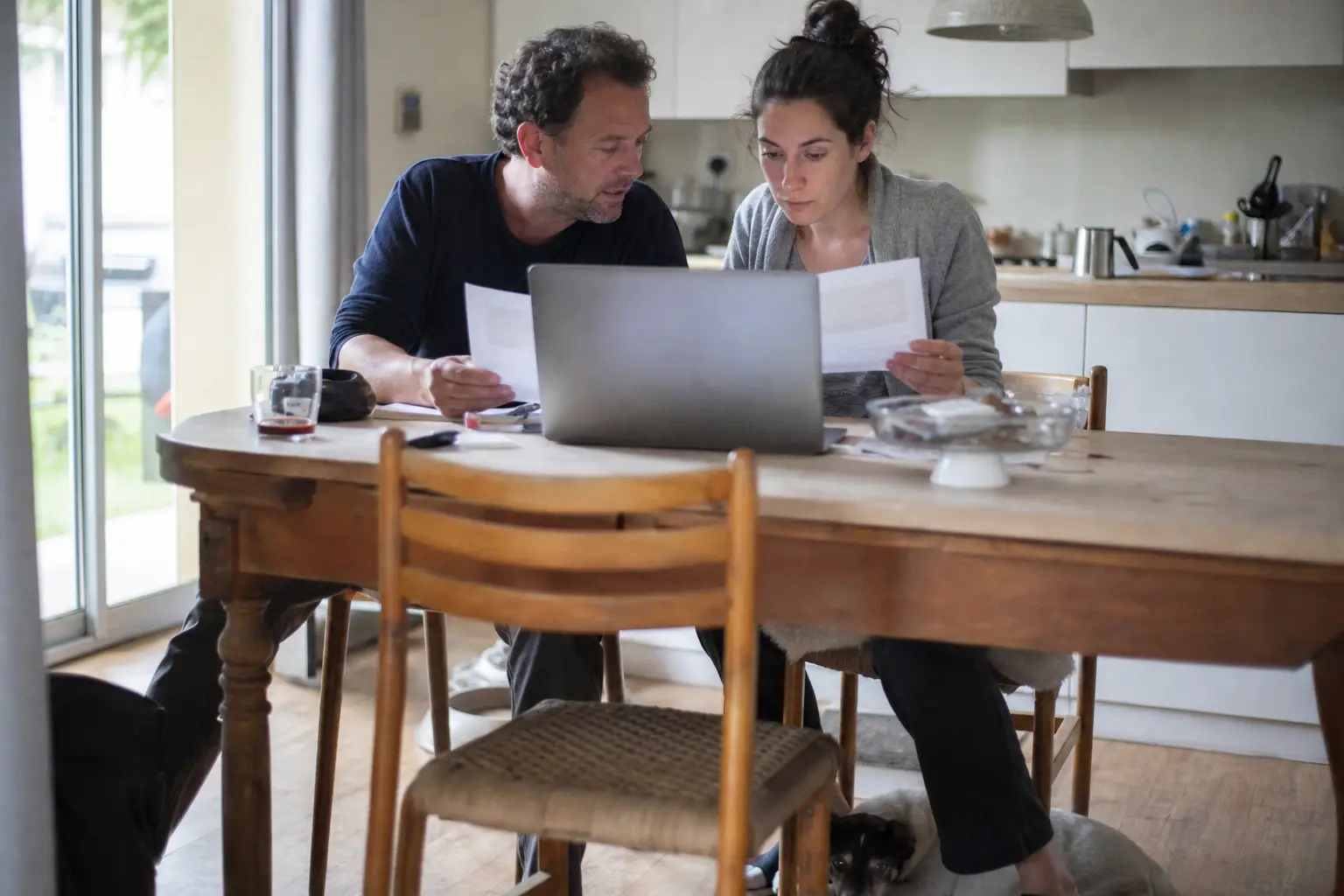 Couple comparant deux documents imprimés à la table de la cuisine, leur chien couché sous la table