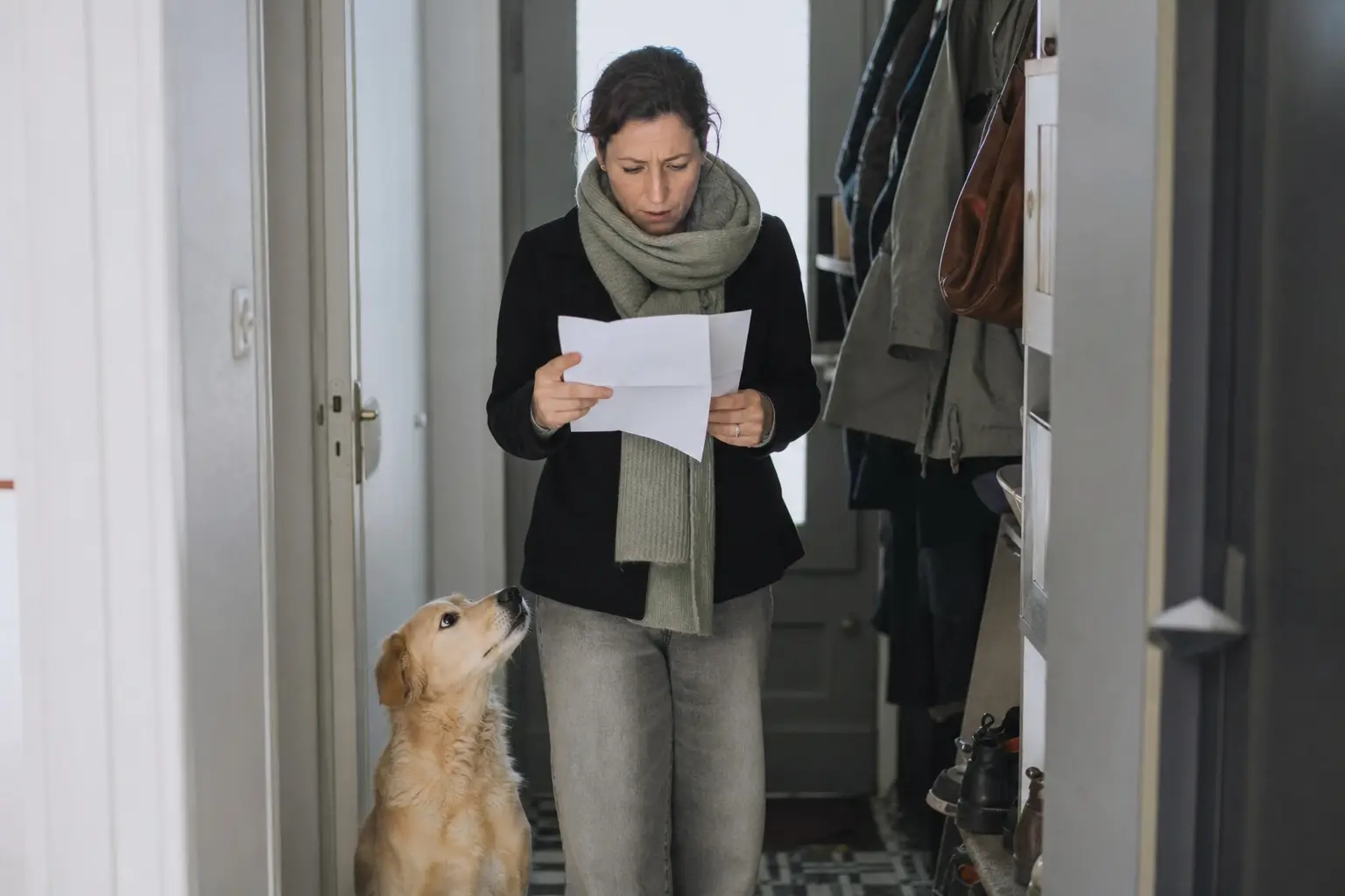 Femme relisant un courrier administratif debout dans son entrée, son chien assis à côté d'elle levant la tête