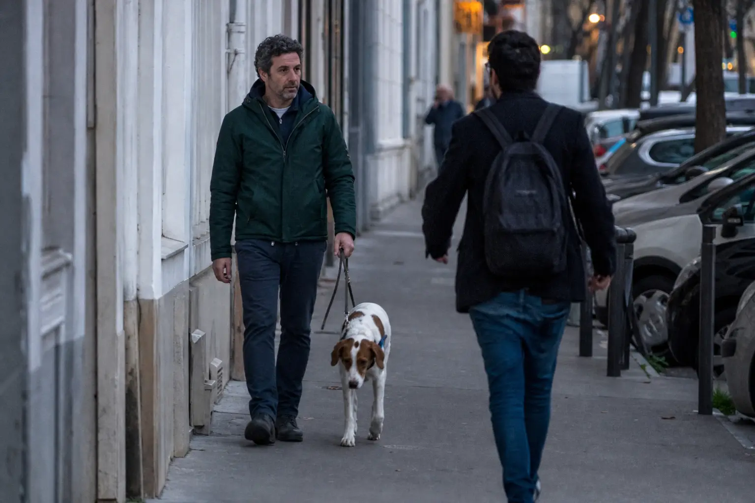 Homme promenant son chien en laisse courte sur un trottoir animé, croisant un autre promeneur